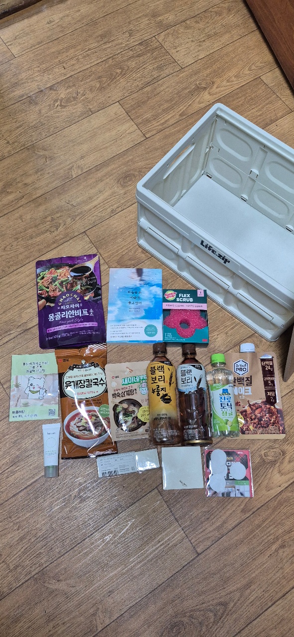 목재 바닥 위에 펼쳐진 식품, 화장품, 생활용품과 흰색 플라스틱 상자