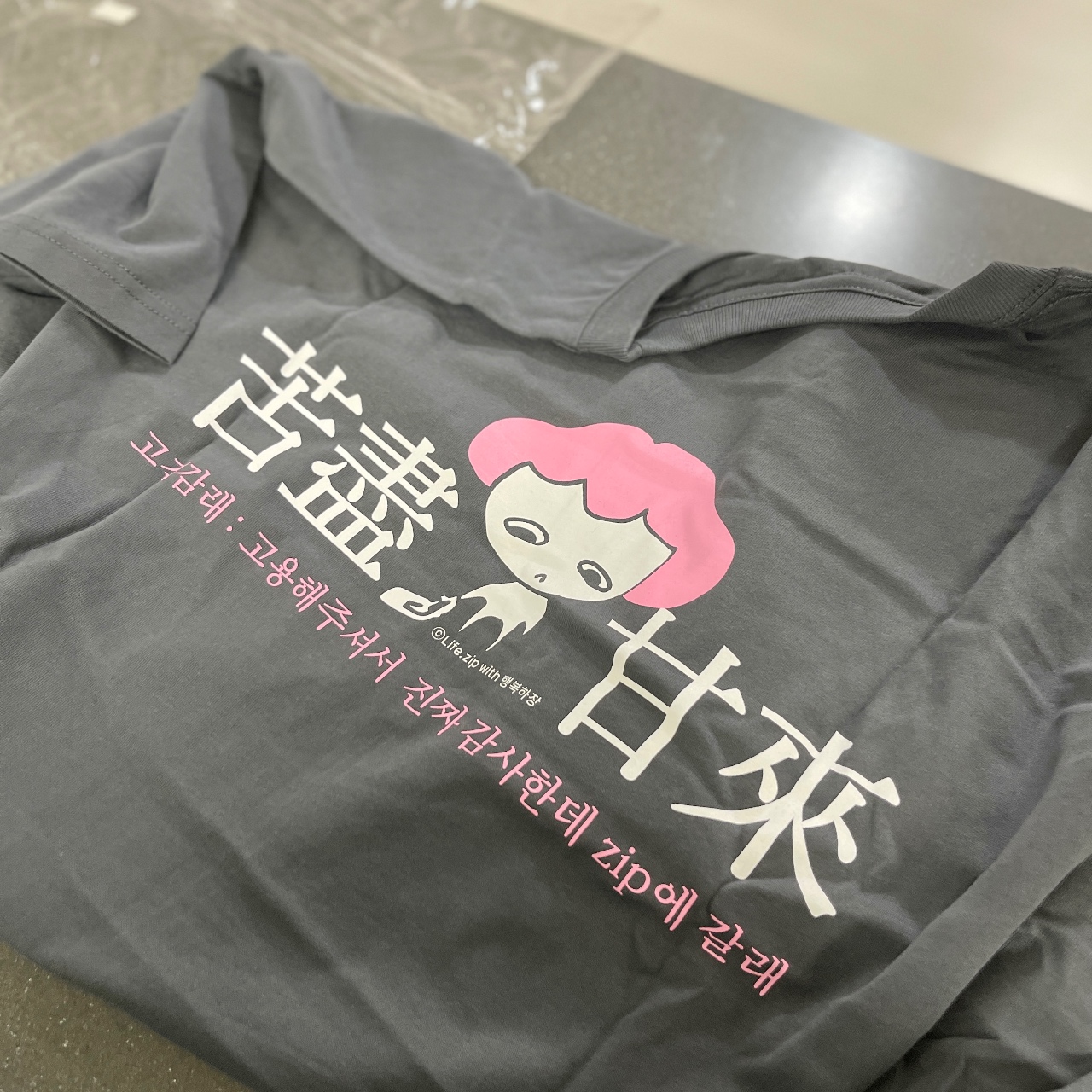 캐릭터 그래픽과 한글·영문 텍스트가 프린트된 회색 반팔 티셔츠