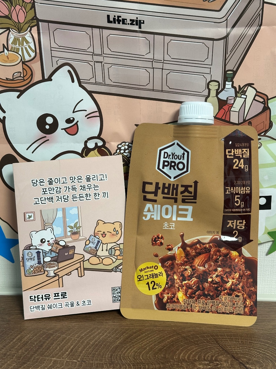 라이프집 로고가 있는 수납장 앞에 전시된 고양이 인형, 단백질 쉐이크, 캐릭터 카드