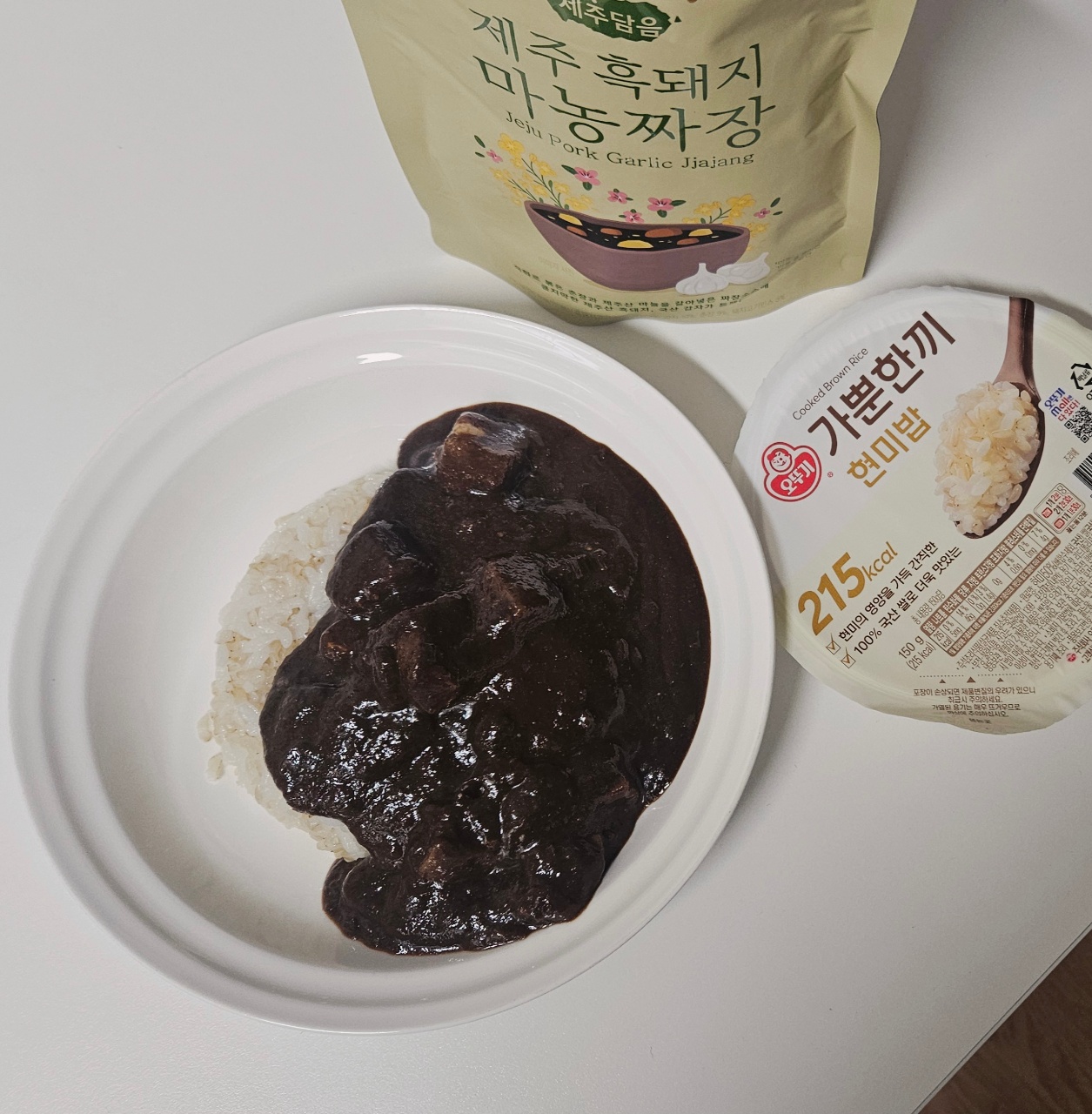 흰 접시에 담긴 검은 소스와 밥, 옆에 놓인 한식 반찬 패키지