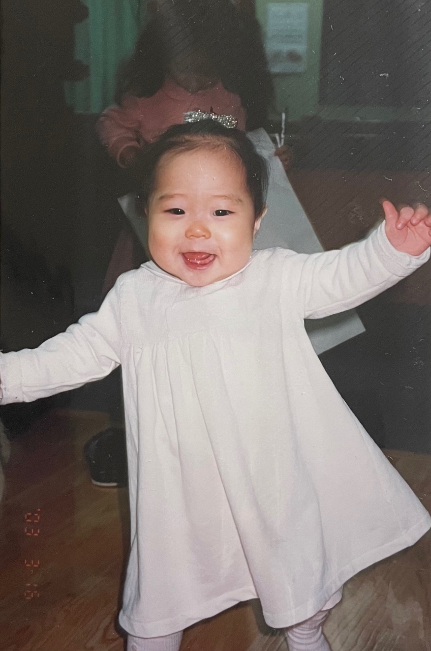 흰 원피스를 입고 머리에 핑크 리본을 쓴 아기가 팔을 벌리고 웃고 있다.